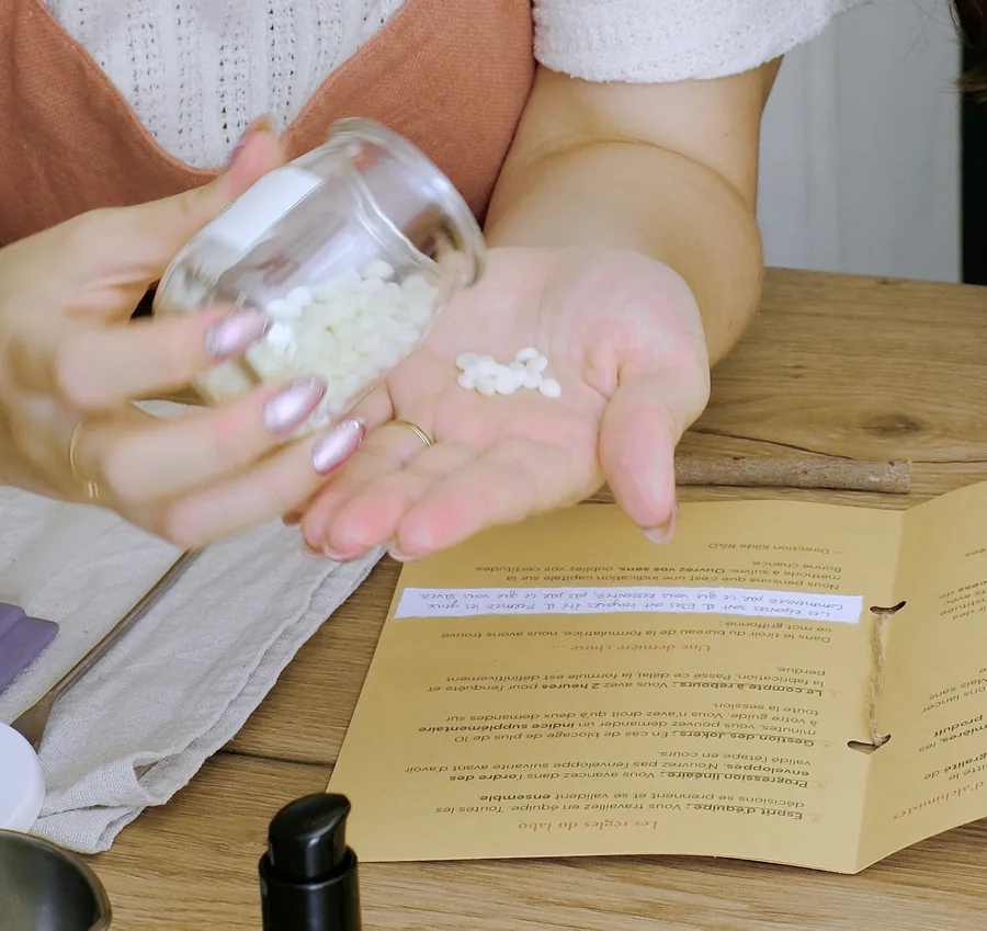 Des paillettes de cire versées d’un bocal dans une paume ouverte, au-dessus du livret de l’atelier